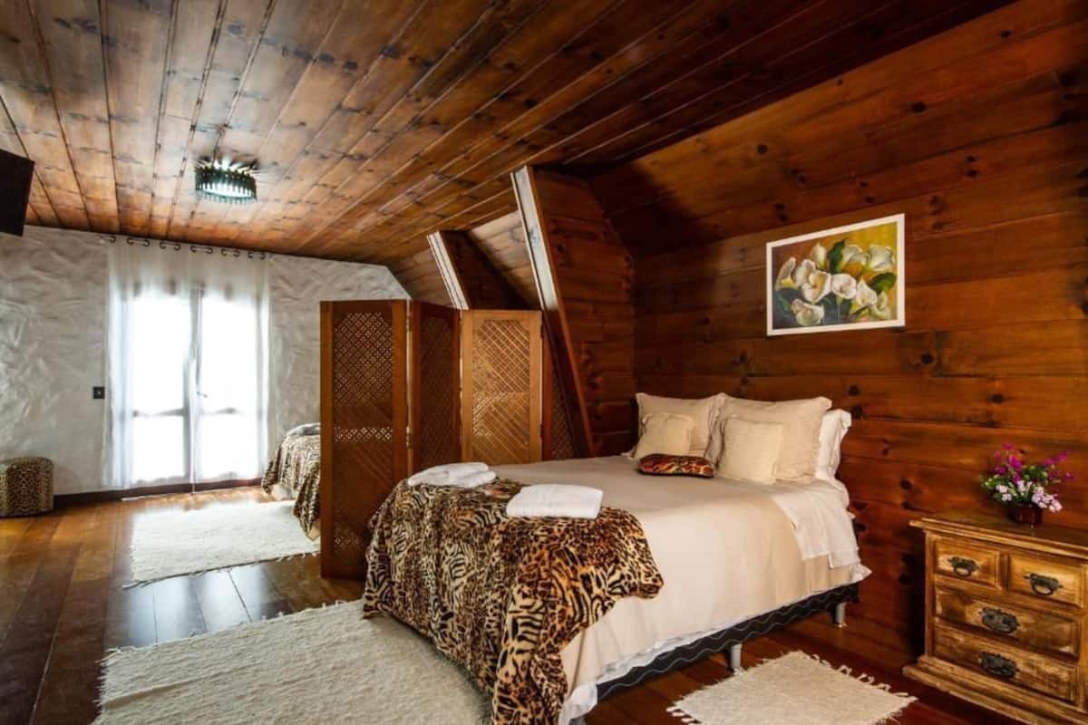 Um quarto de madeira com uma cama, um aparador de madeira, um quadro e uma janela com cortinas.