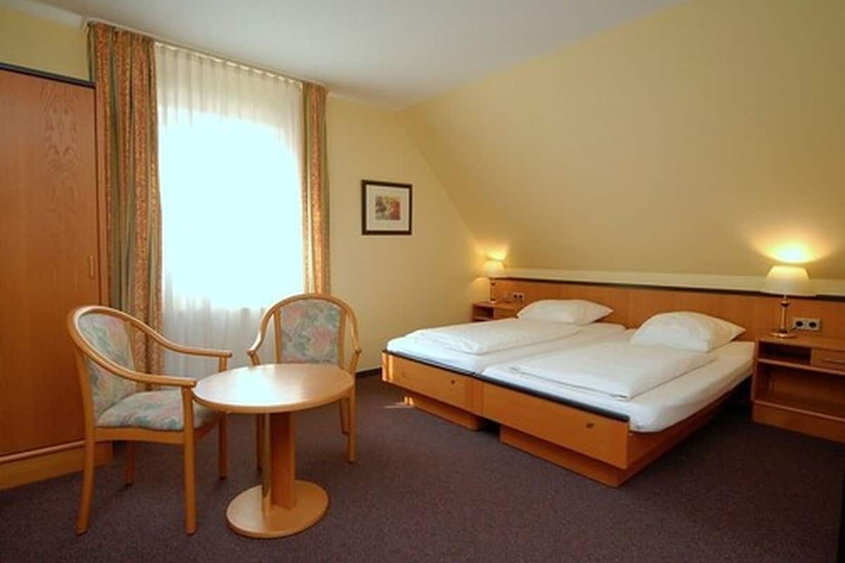 A hotel room with a bed, two chairs, a small table, a nightstand, and a lamp.