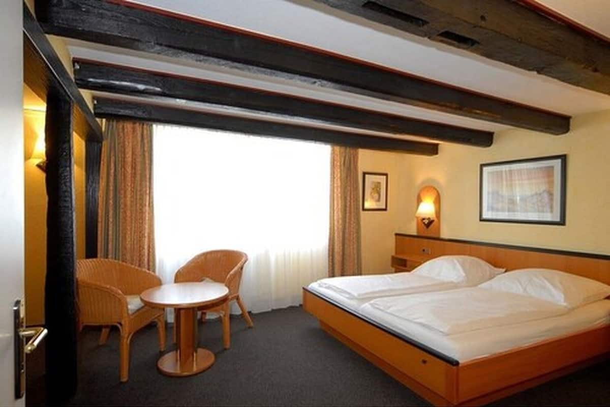 A hotel room with a bed, a small table, two chairs, a window with curtains, and a wall-mounted lamp.