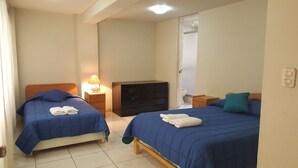Un dormitorio con dos camas, cada una con colcha y almohadas azules, un aparador de madera, una mesita de noche con lámpara, y una puerta que da a un baño.
