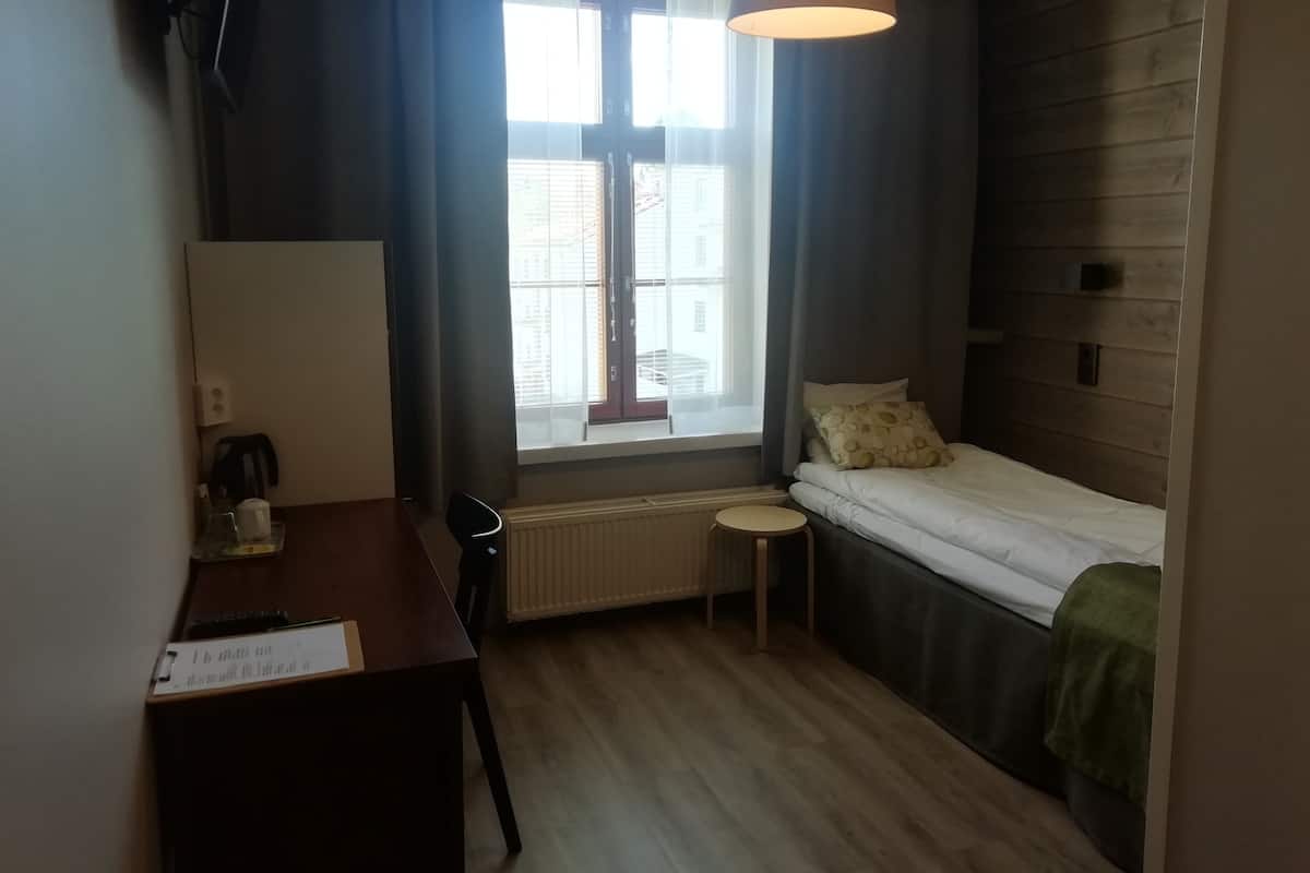 A small, well-lit room with a bed, a desk, a chair, a window with curtains, and a radiator.
