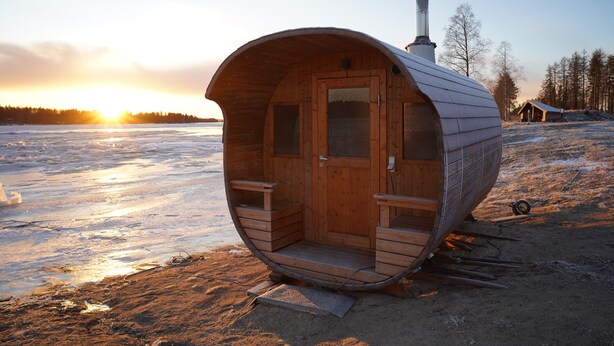 Een houten sauna met een deur en twee bankjes, gelegen in een besneeuwd landschap met een zonsondergang op de achtergrond.