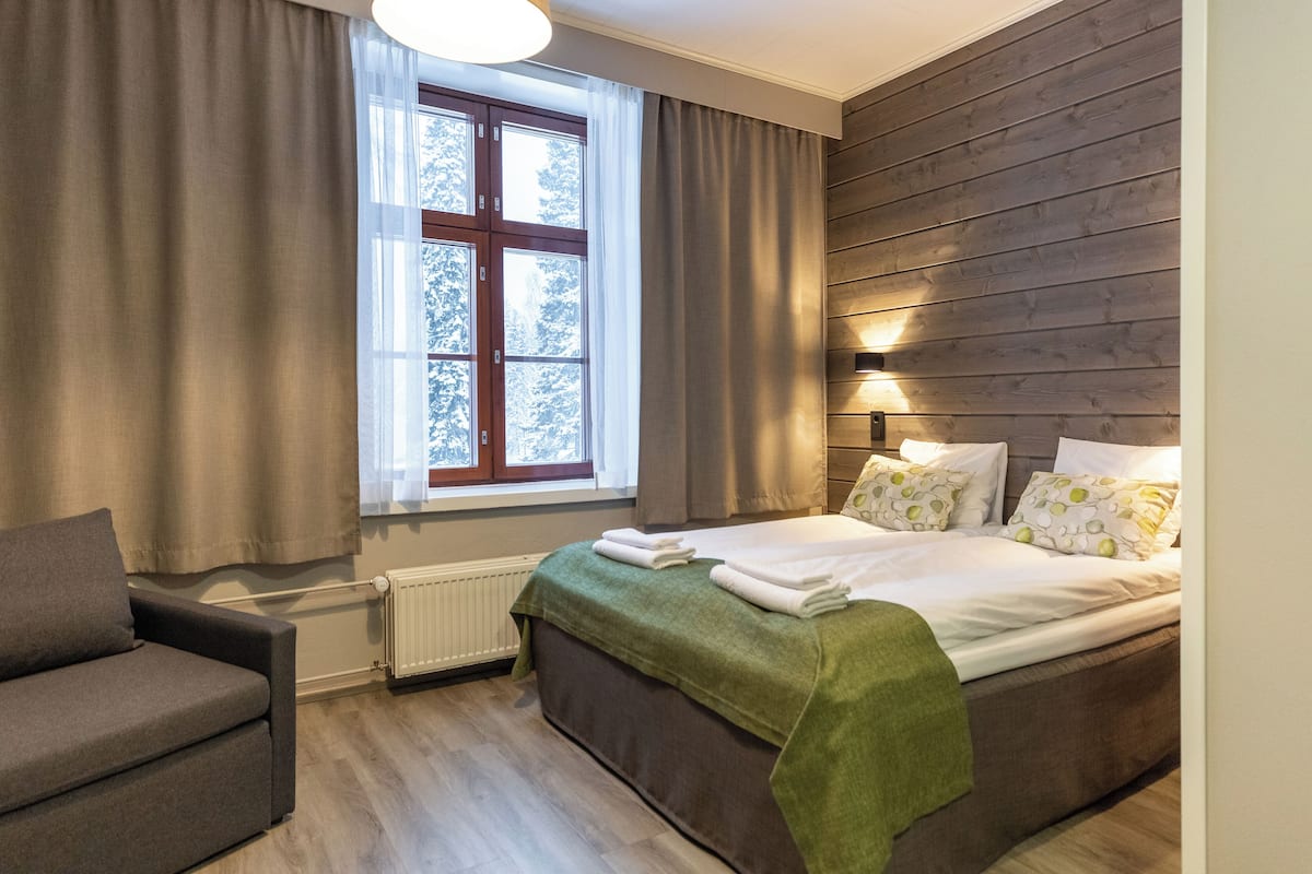 A bedroom with a bed, a window with curtains, a radiator, and a wall with wooden paneling.