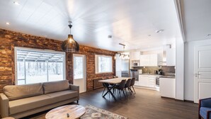Een moderne woonkamer met een bank, salontafel en eethoek. Er zijn grote ramen, een keuken met witte kasten en een accentmuur van hout.