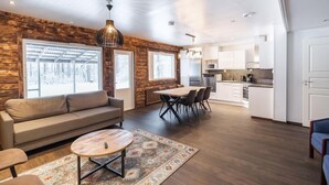 Een moderne woonkamer met een bank, salontafel en een eethoek. Er zijn grote ramen, een keuken met witte kasten en een houten accentmuur.