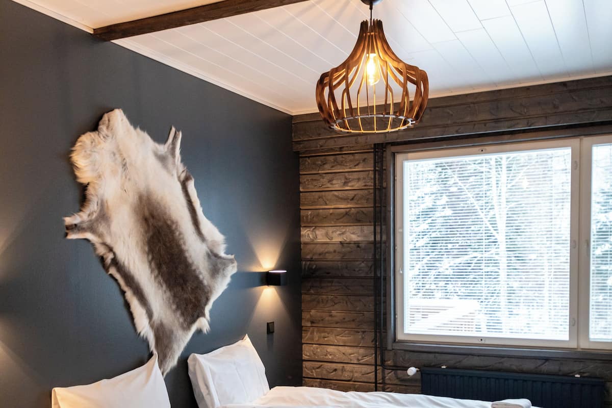 A modern hotel room with a large bed, a blue chair, a wall-mounted light fixture, and a window with a view of trees.