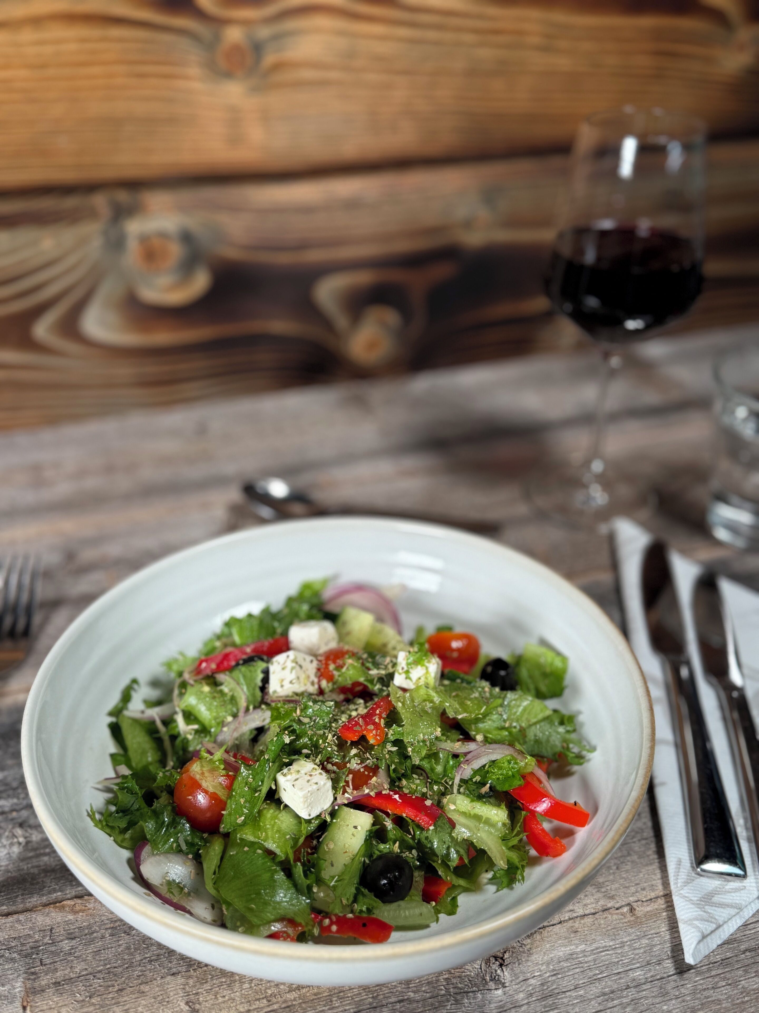 Ensalada con diversas verduras y queso, acompañada de una copa de vino tinto y cubiertos sobre una mesa de madera.