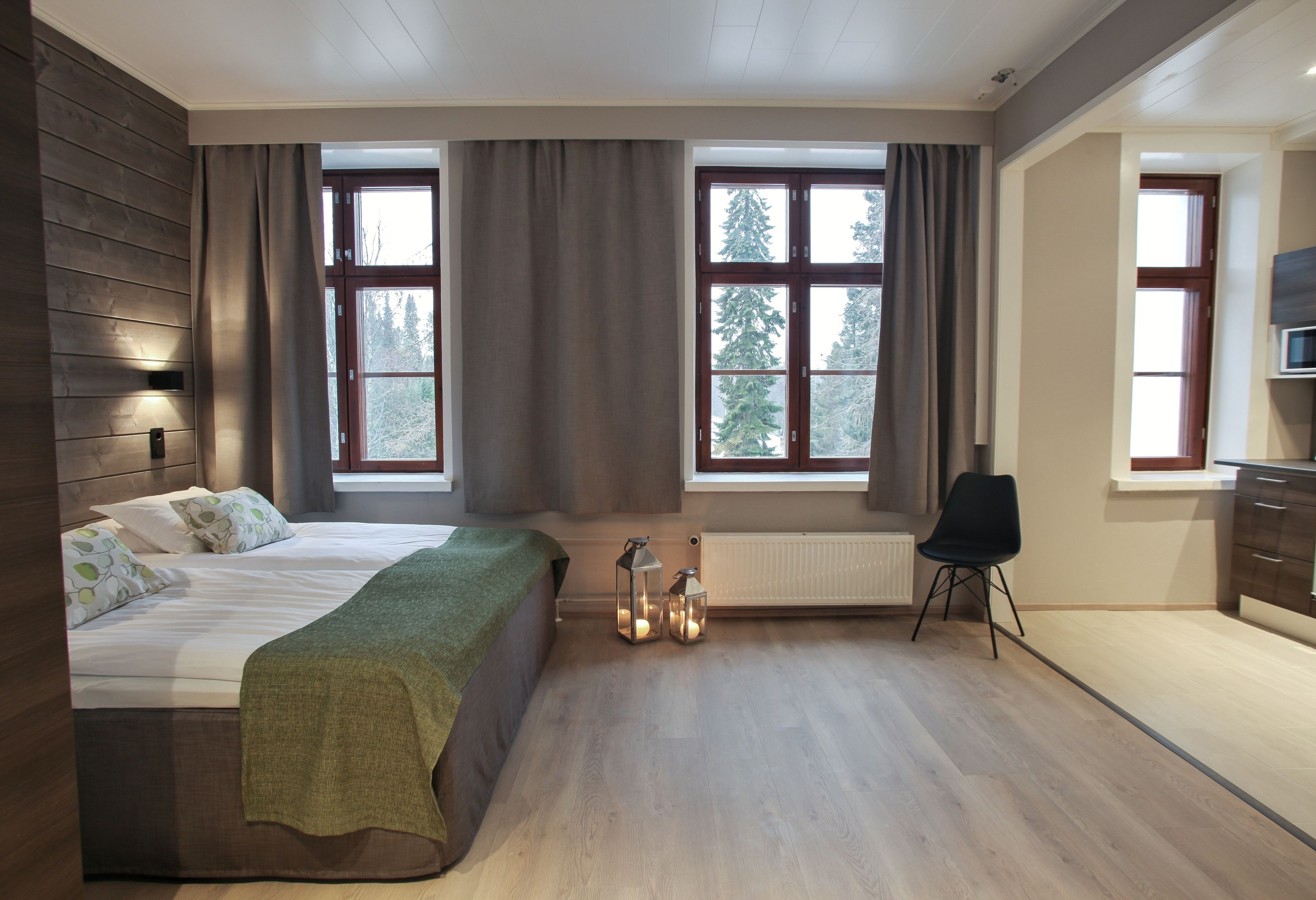 Un dormitorio moderno con una cama grande, un escritorio y una silla. Hay dos ventanas con cortinas, un radiador y una lámpara de pared.