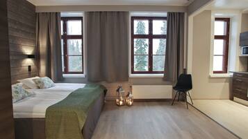 Un dormitorio moderno con una cama grande, un escritorio y una silla. Hay dos ventanas con cortinas, un radiador y una lámpara de pared.