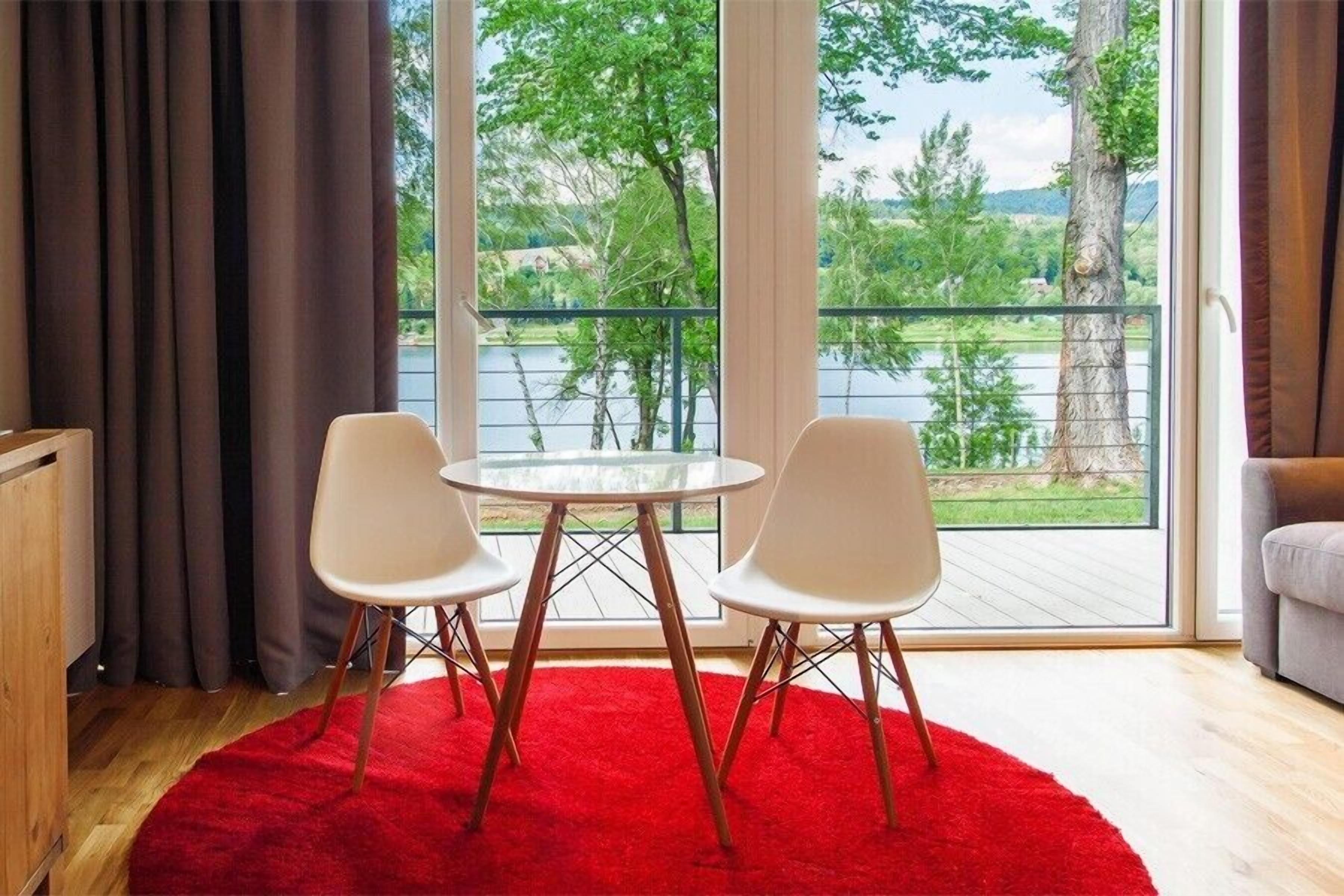 A room with a large window, a round table with two chairs, a red rug, and a view of a lake and trees.