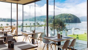 A restaurant with large windows offering a view of a lake and forested hills. Tables are arranged with chairs, and there are potted plants on the tables. - Lemon Resort Spa (Grodek Nad Dunajcem)