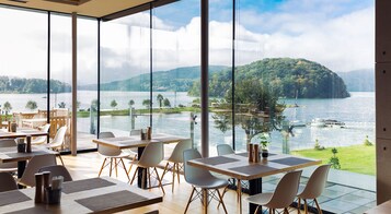 Restaurante com grandes janelas oferecendo vista para um lago e colinas arborizadas. Mesas dispostas com cadeiras, e plantas em vasos sobre elas.