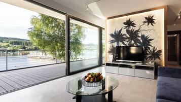 A modern living room with a large glass table, a flat-screen TV, and a view of a river and greenery.