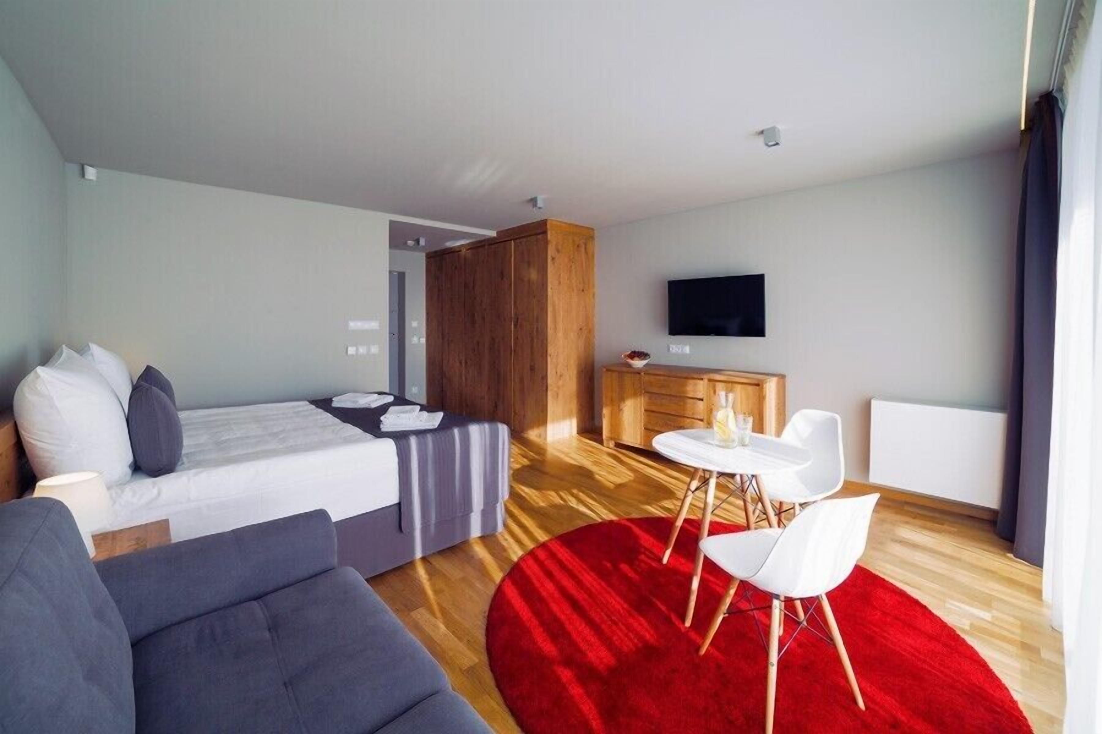 A modern hotel room with a bed, a sofa, a dining table with chairs, a TV, and a red rug.