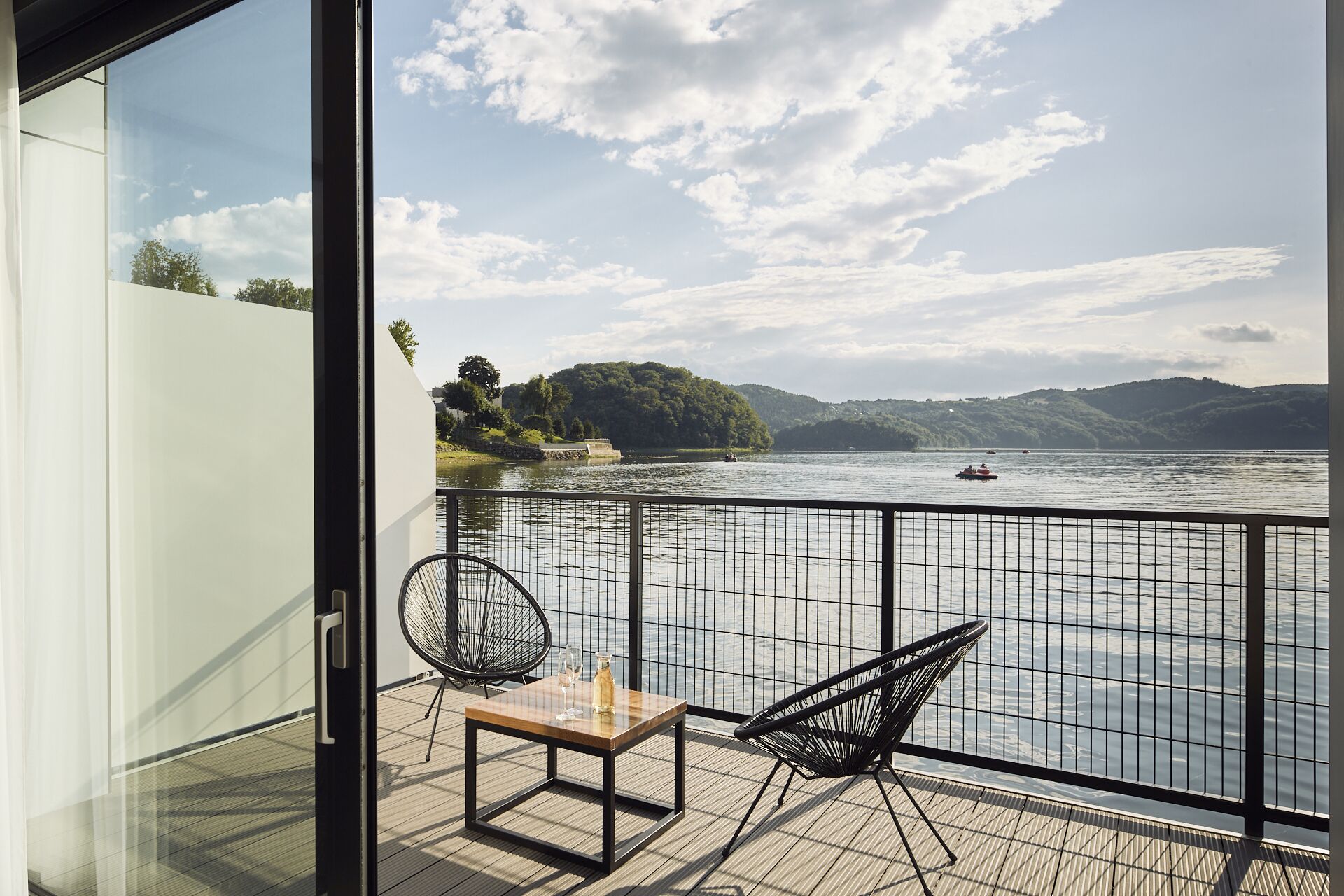 Un balcone con un tavolo in legno e due sedie, affacciato su un fiume con barche e vegetazione sullo sfondo.