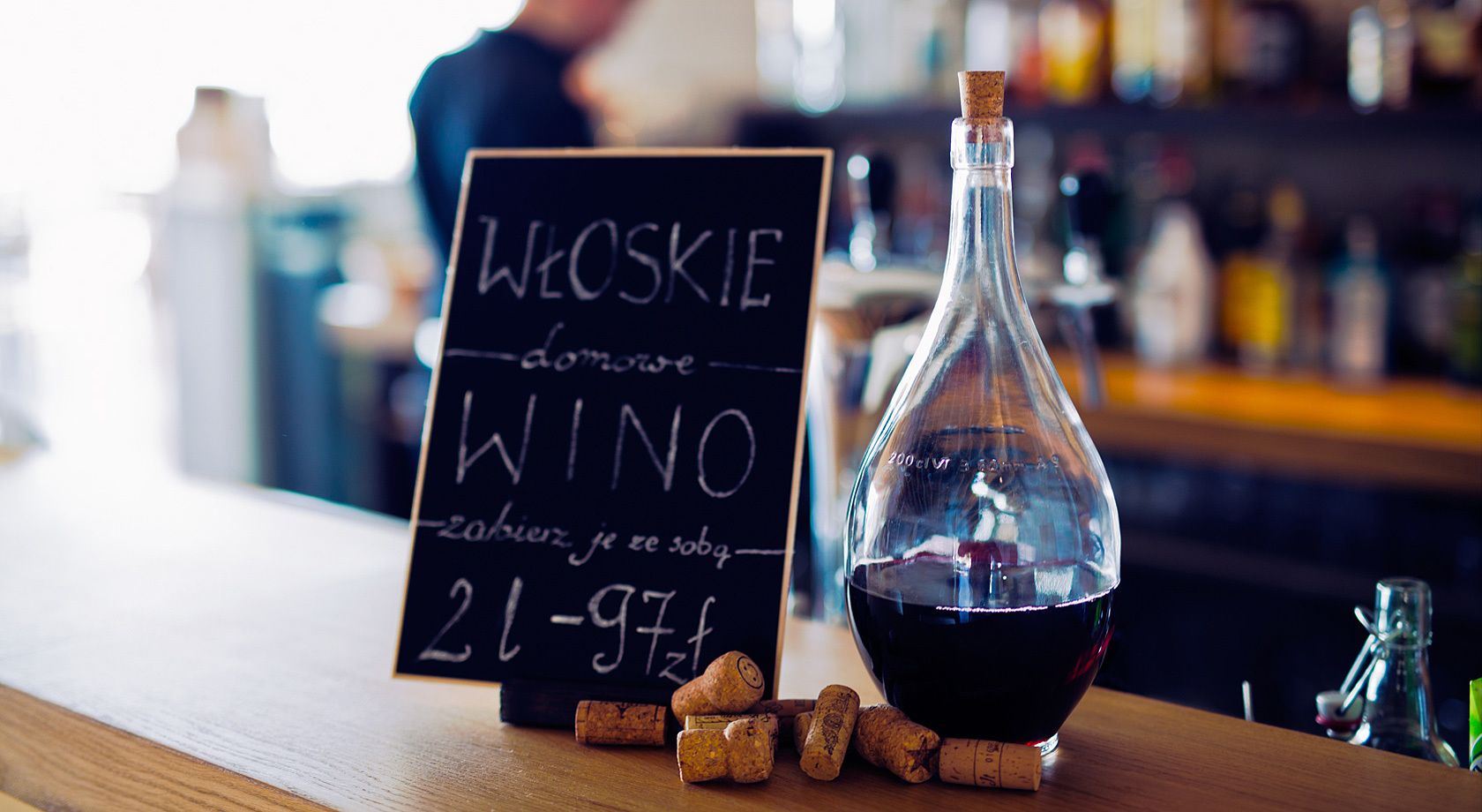 Uma garrafa de vinho tinto, rolhas e um menu em lousa com o texto "WOSKIE DOMOWE WINO".