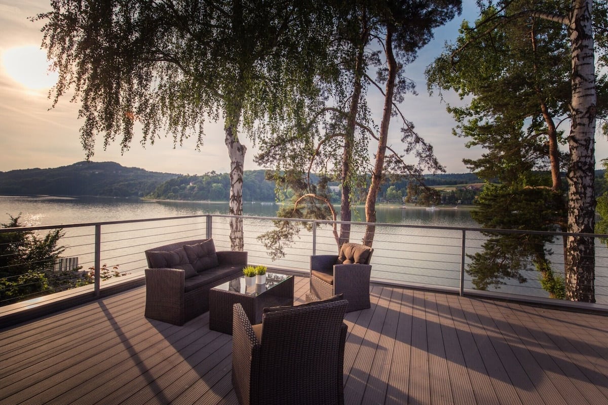 A deck with wicker furniture and a glass table, overlooking a lake with trees and hills in the background.