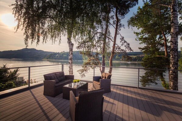 A deck with wicker furniture and a glass table, overlooking a lake with trees and hills in the background. - Lemon Resort Spa (Grodek Nad Dunajcem)