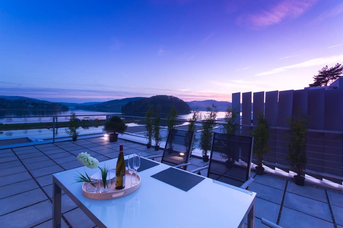 A terrace with a table set for a meal, overlooking a river and hills.