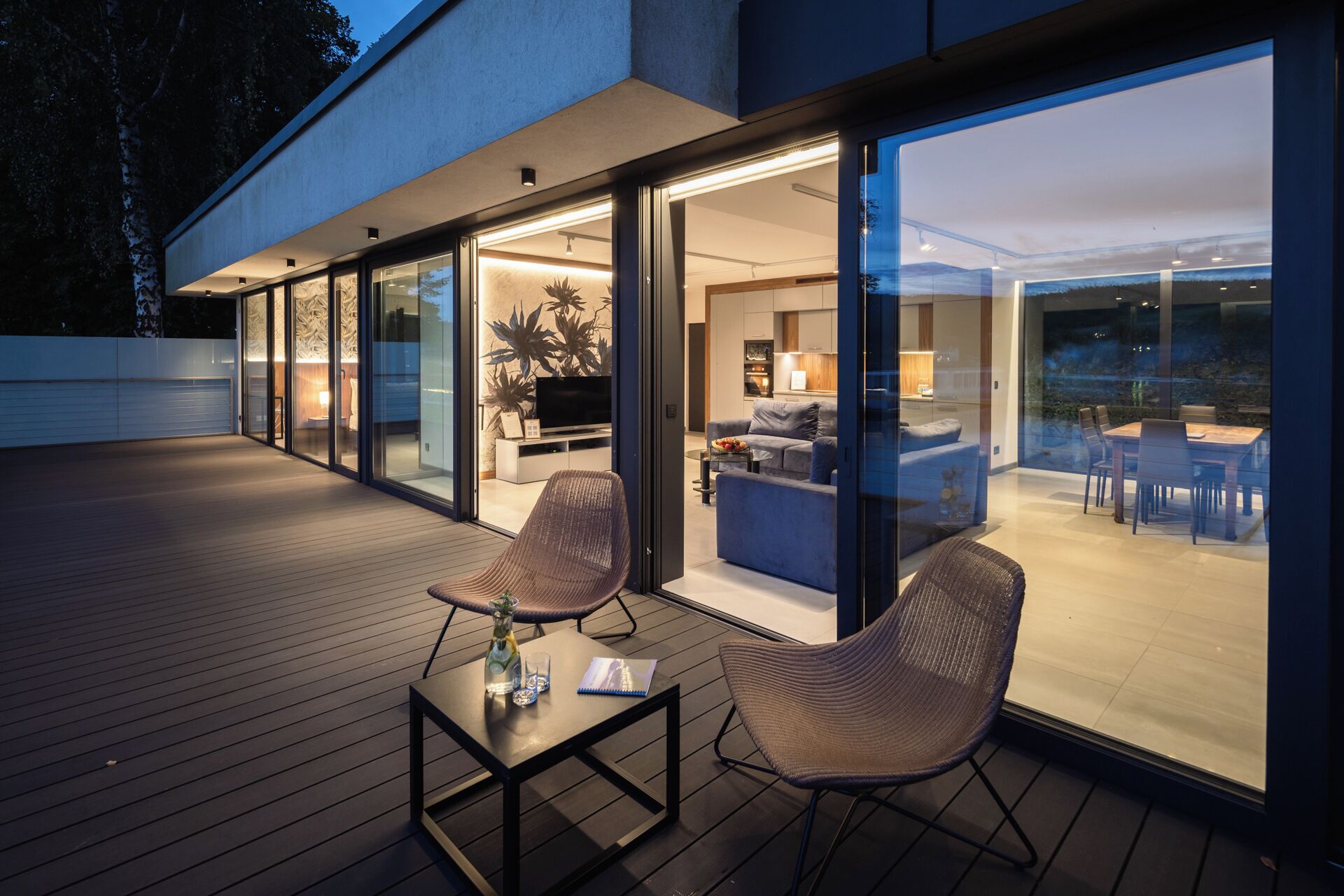 An outdoor living area with a glass-enclosed space, a dining table, and two wicker chairs.
