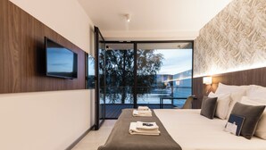 A modern hotel room with a large bed, a flat-screen TV mounted on the wall, a balcony with a view of a lake, and a patterned wall. - Lemon Resort Spa (Grodek Nad Dunajcem)