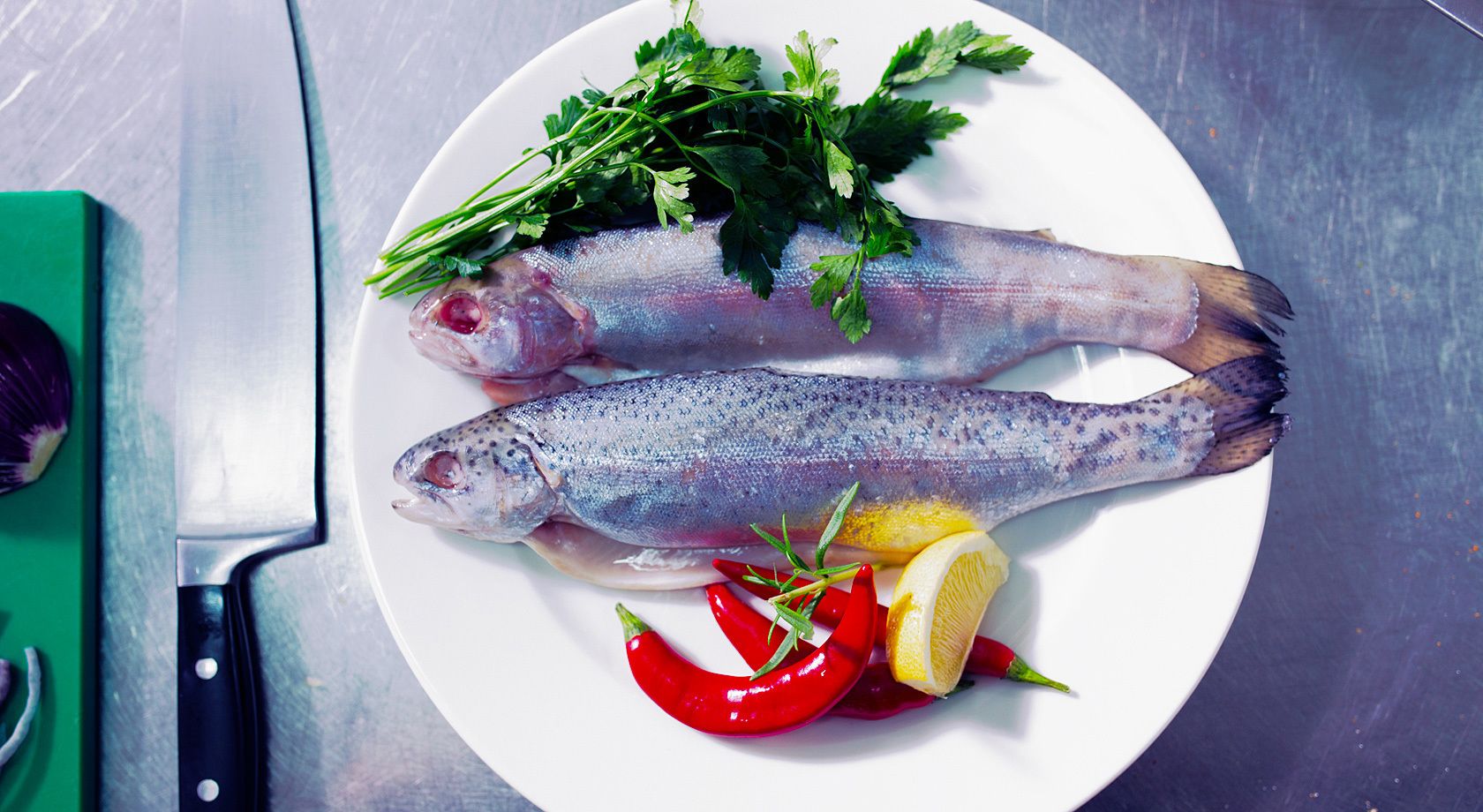 Dos pescados enteros sobre un plato blanco con perejil, limón y chiles rojos.