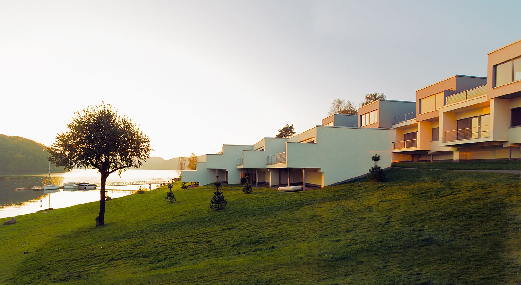 Un edificio moderno con techo plano y un lado inclinado, ubicado en una colina cubierta de césped con vista a un cuerpo de agua.