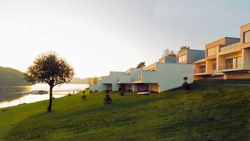 Un edificio moderno con techo plano y un lado inclinado, ubicado en una colina cubierta de césped con vista a un cuerpo de agua.