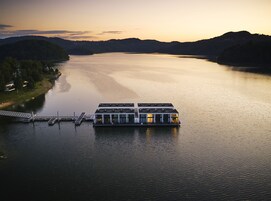 Uma casa barca moderna flutuando em um lago calmo, com um cais e cercada por colinas.