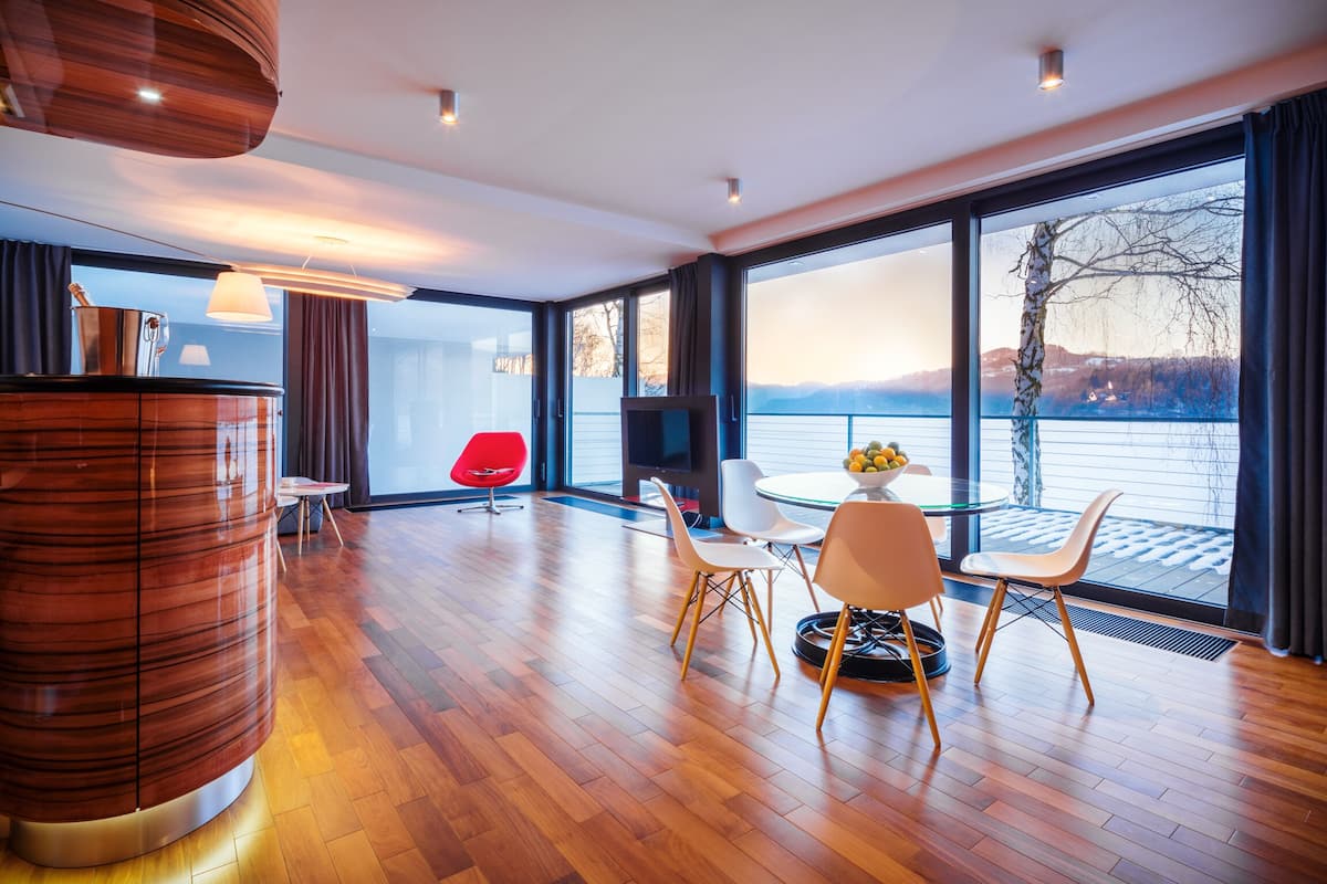 A modern room with a wooden floor, a red chair, a round table with chairs, a TV, and large windows with a view of a lake and mountains.