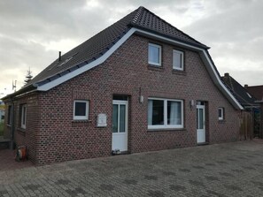 Une maison à deux étages, construite en briques, avec un toit à pignon, deux fenêtres au rez-de-chaussée et un petit porche.