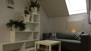 Ein modernes Wohnzimmer mit einem grauen Sofa, einem weißen Couchtisch und einem Streifenmuster-Teppich. Weiße Regale mit Blumentöpfen und ein Oberlicht sind ebenfalls vorhanden.