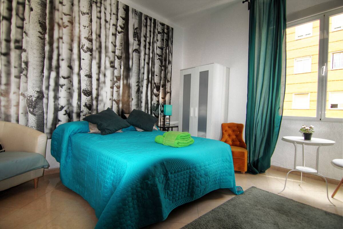 A bedroom with a bed, a sofa, a chair, a wardrobe, and a small table with a plant.