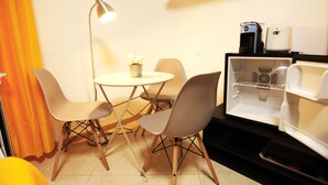 Ett litet rum med ett litet bord och fyra stolar, en golvlampa, ett kylskåp med en kaffebryggare och en krukväxt.