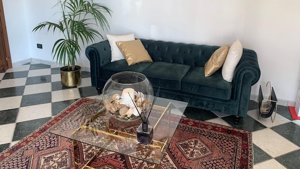 Uma sala de estar com um sofá verde escuro, um tapete estampado, uma planta em vaso e uma mesa de centro de vidro com objetos decorativos.