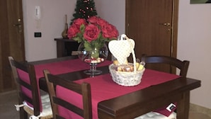 Uma sala de jantar com uma mesa de madeira, toalha de mesa vermelha e quatro cadeiras. Uma árvore de Natal e um vaso com rosas vermelhas estão sobre a mesa.