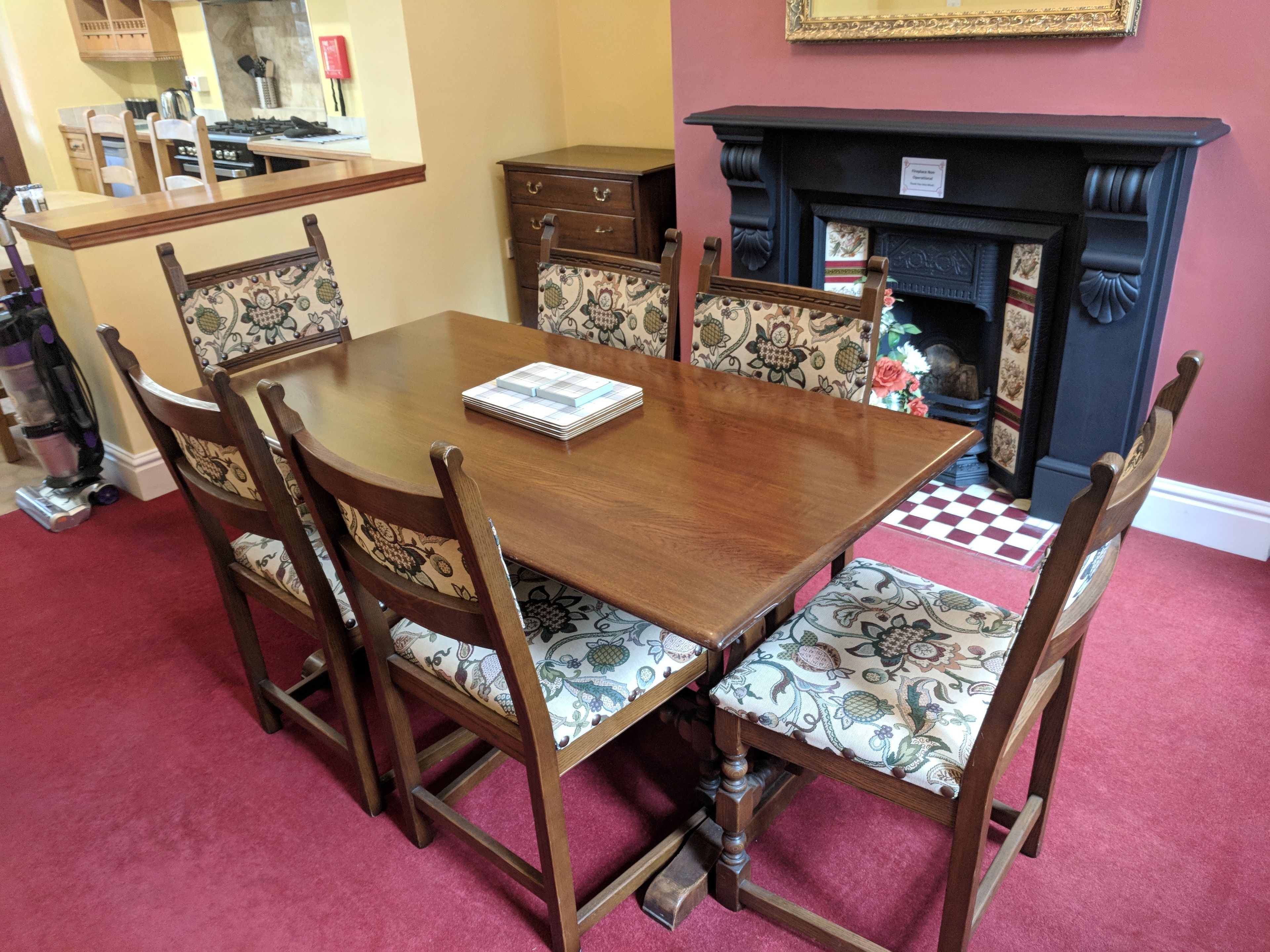 A dining room with a wooden table and chairs, a fireplace, and a kitchen area in the background.