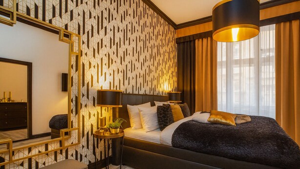 Quarto de hotel moderno com parede estampada em padrão geométrico, uma cama grande com várias almofadas, mesa de cabeceira com abajur e uma janela ampla com cortinas.