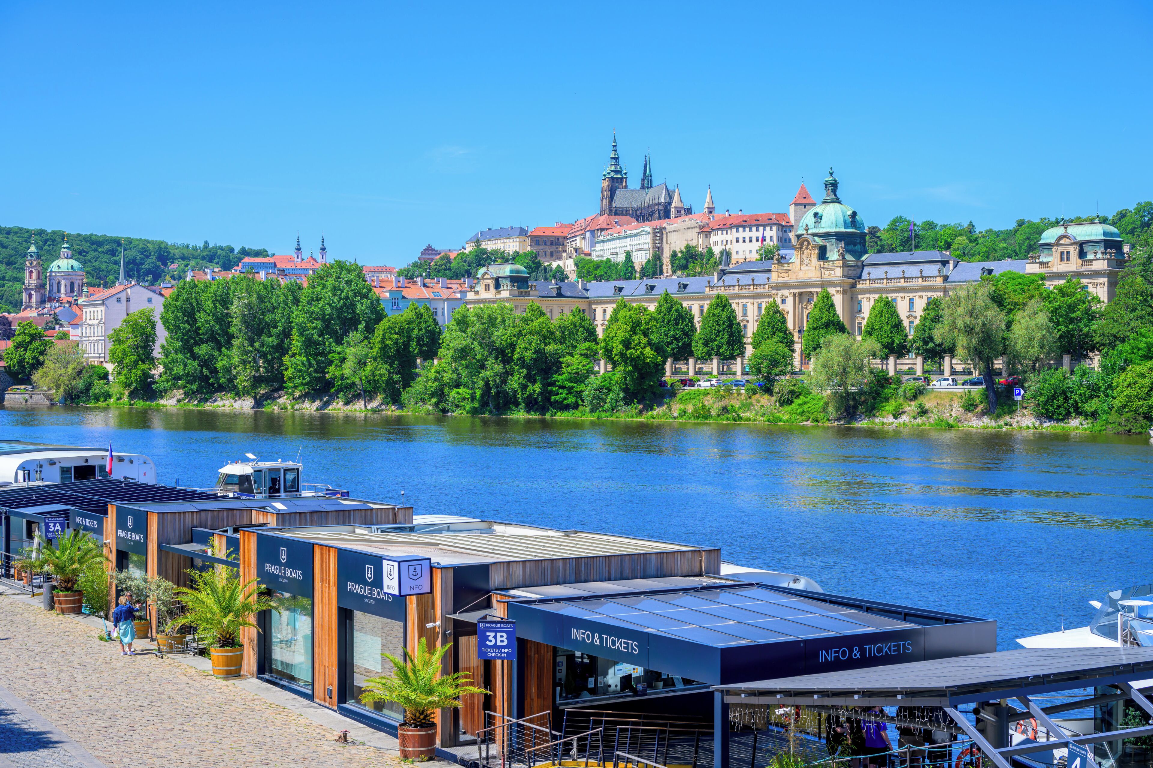 A riverside view with a historic building complex in the background and a modern boat terminal in the foreground.