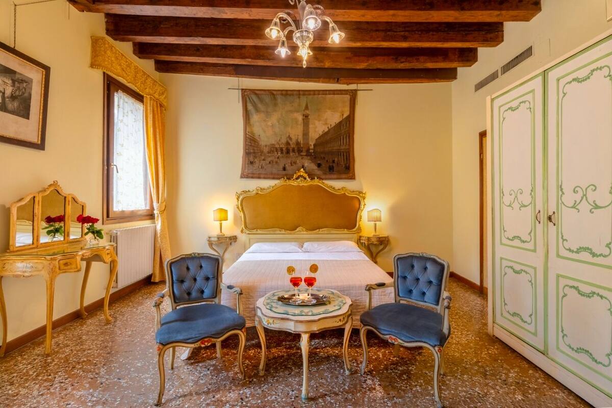 A room with a bed, two chairs, a table with glasses, a mirror, a painting, and a wardrobe.