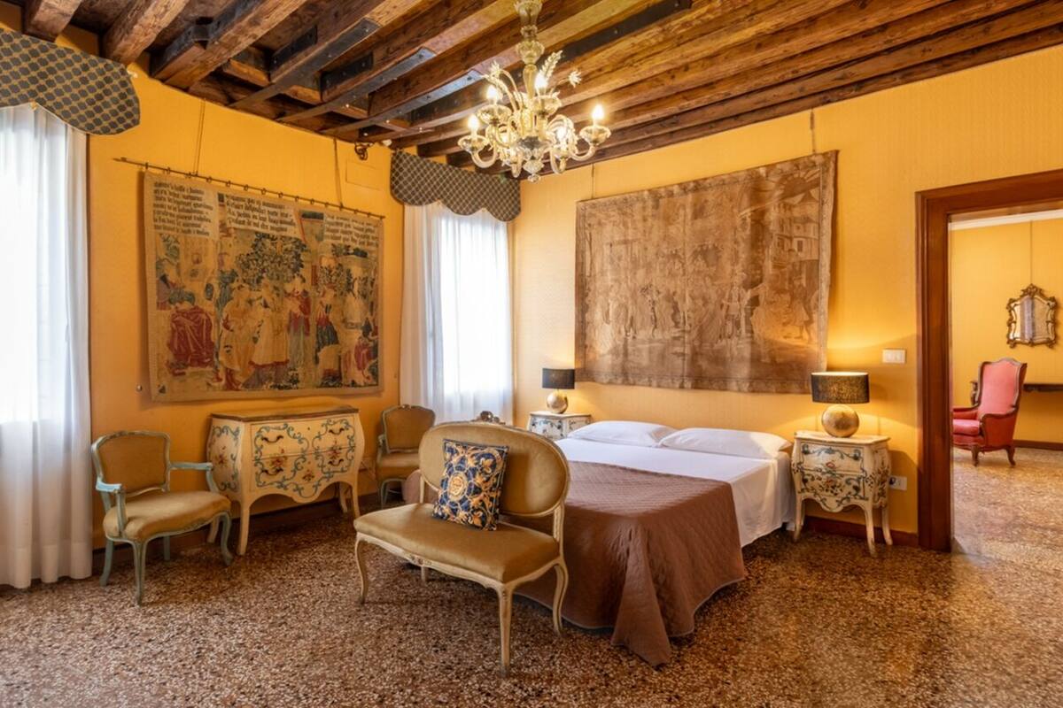 A hotel room with a bed, a desk, a chair, a chandelier, and artwork on the walls.