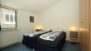 A bedroom with two beds, a window, a radiator, a wooden wardrobe, and a small bedside table with a lamp.