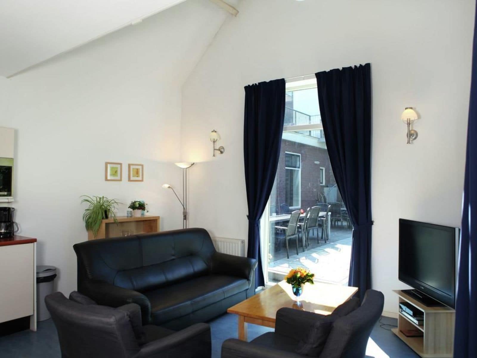 Una sala de estar con un sofá negro, dos sillones, una mesa de centro, un televisor y un ventanal con cortinas.