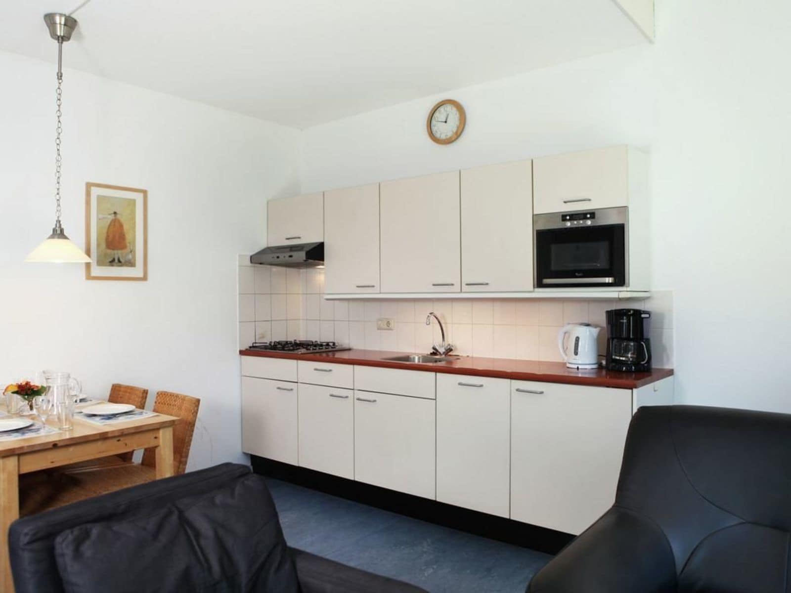 A compact kitchen with white cabinets, a stove, a microwave, and a coffee maker. There is a dining table with chairs and a clock on the wall.