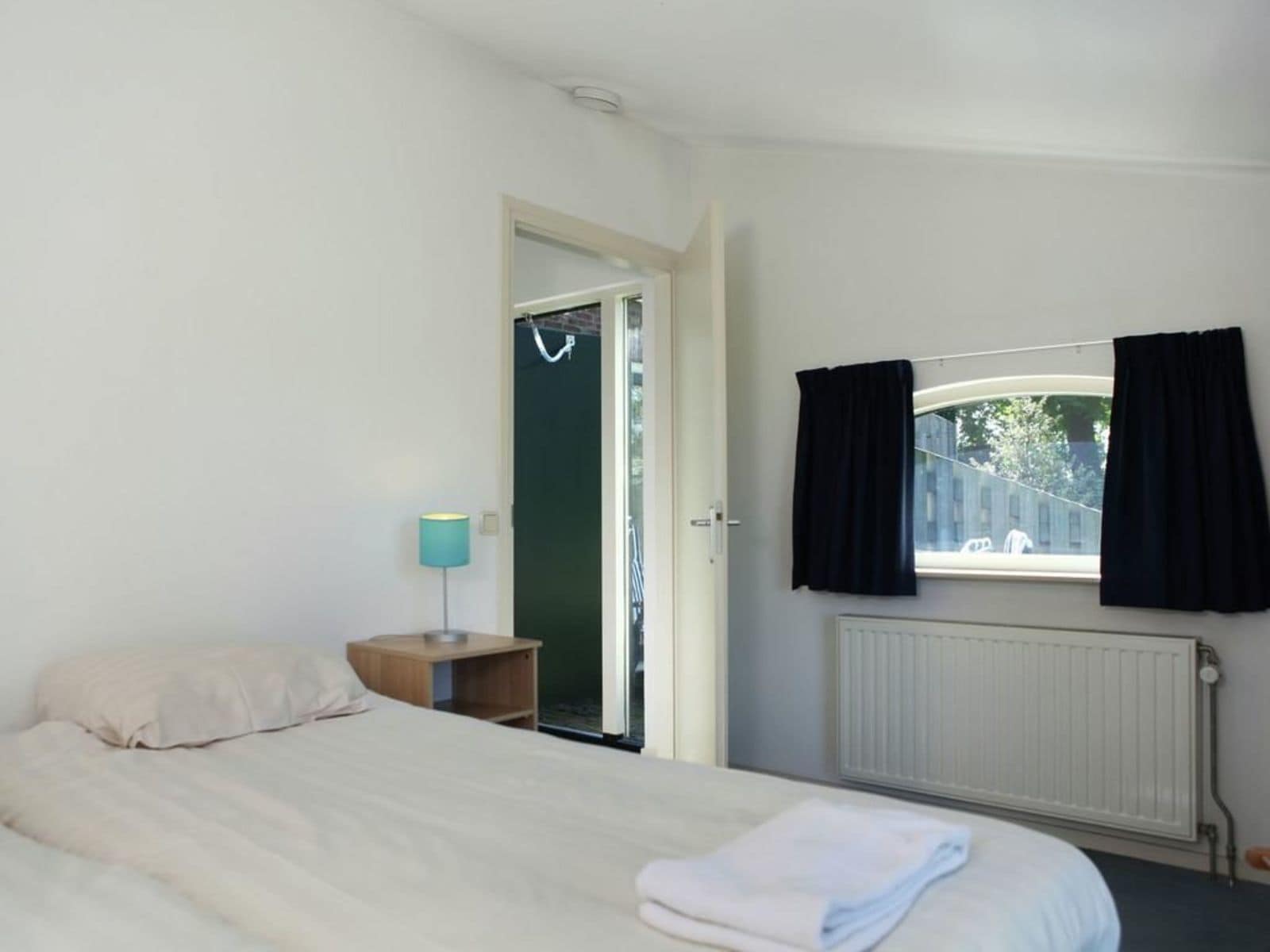 Et soveværelse med seng, et natbord, en lampe, et vindue med gardiner og en radiator.