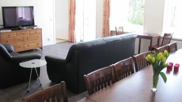 A living room with a black sofa, wooden chairs, a dining table, a small TV on a wooden cabinet, and a vase with yellow flowers.