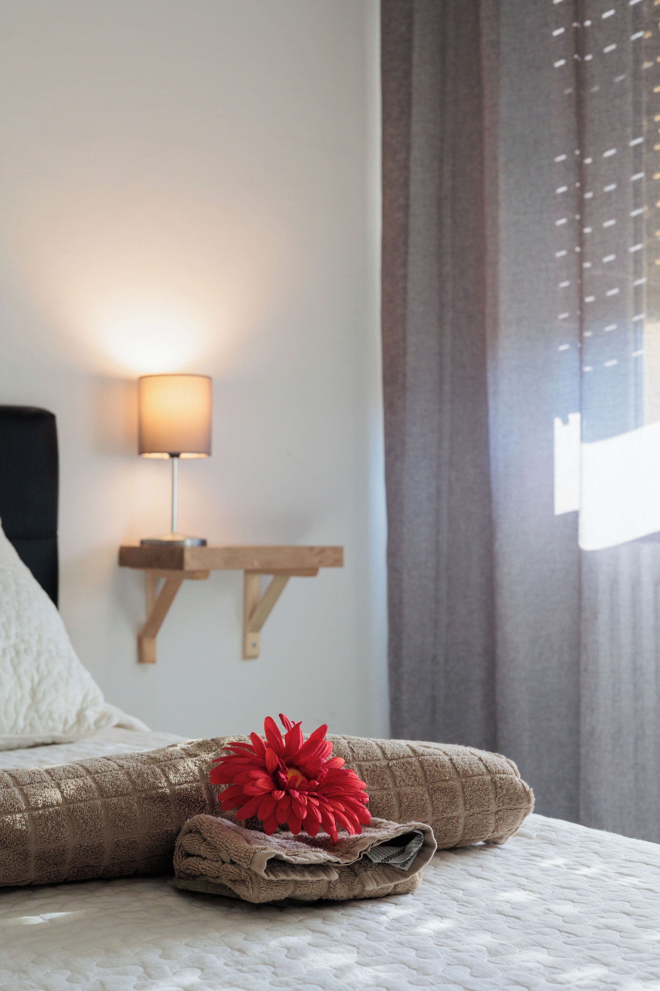 Un lit bien fait, avec une serviette pliée et une fleur rouge, une lampe de chevet et une table de nuit en bois, le tout placé contre un mur avec une fenêtre et des rideaux.