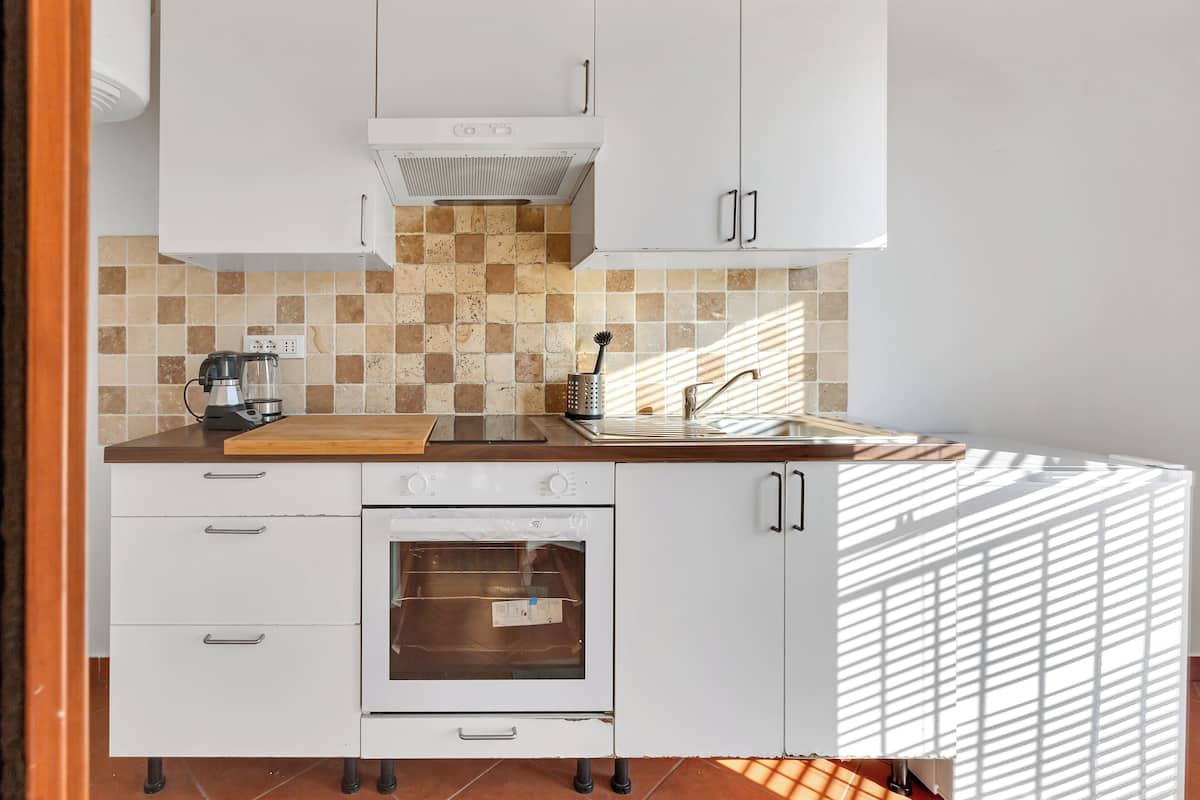 Une cuisine avec des meubles blancs, un pare-feu en carreaux, une machine à café, un évier et une fenêtre qui projette des ombres.