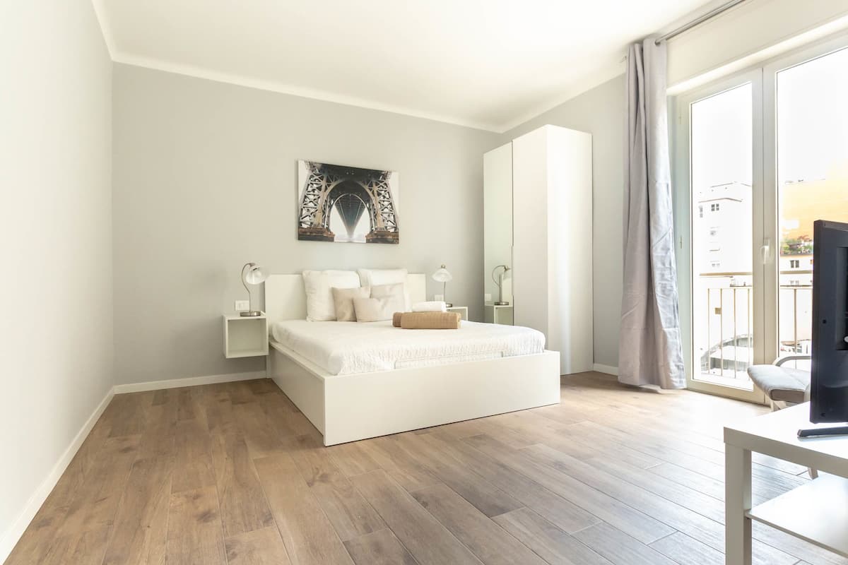 Une chambre moderne avec un grand lit, une table de chevet, une commode, un téléviseur et une fenêtre avec des rideaux.