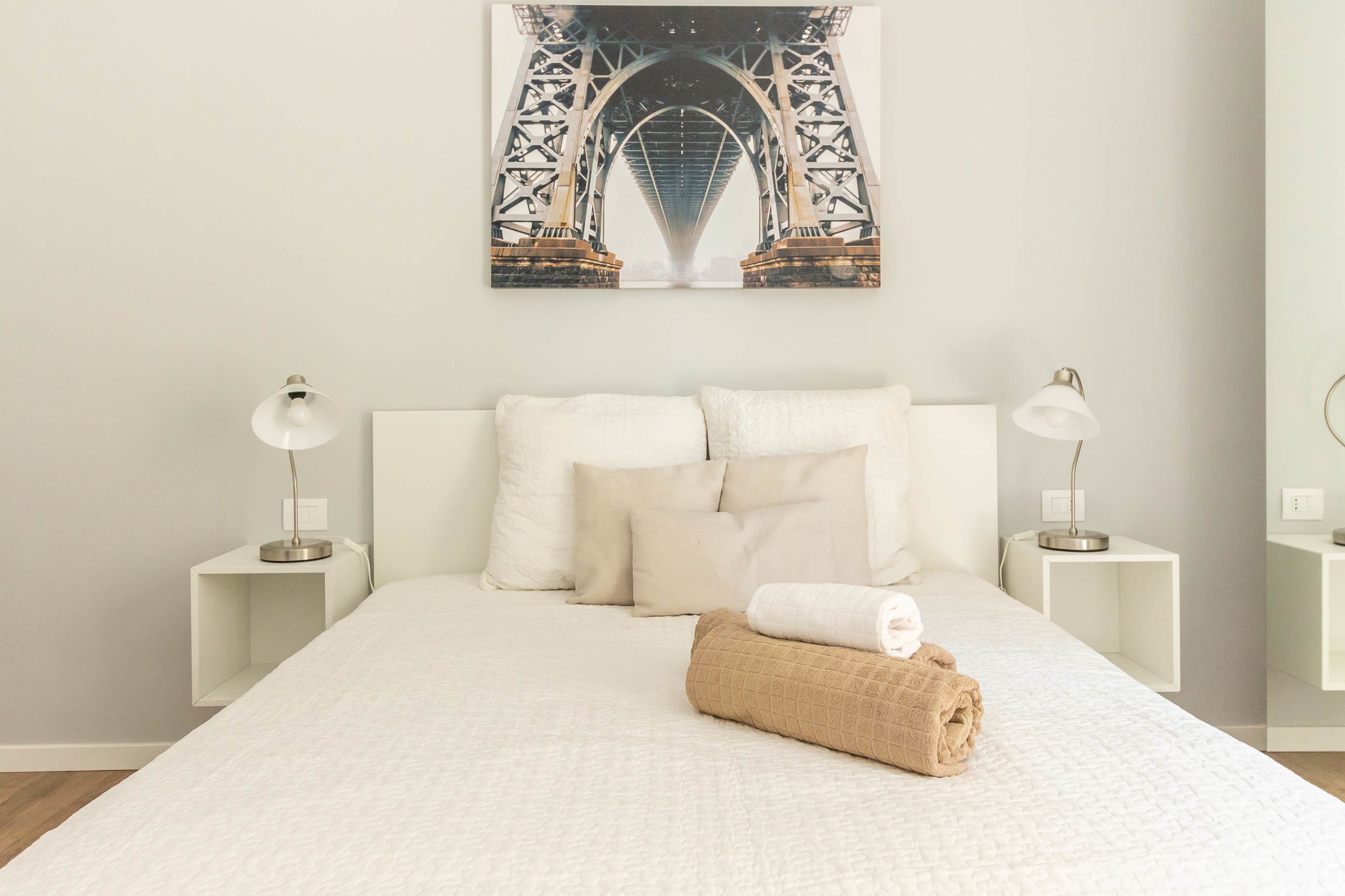 A hotel room with a large bed, white bedding, two bedside tables with lamps, and a framed picture of a bridge on the wall.
